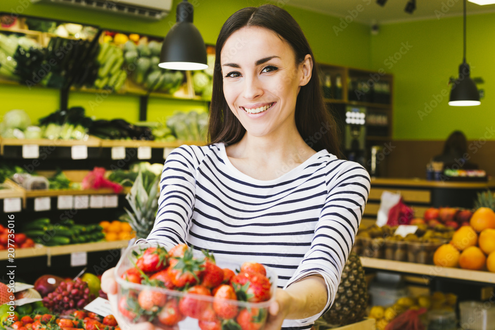 Beautiful young smiling woman holding a box with fresh strawberries and showing at the camera in a supermarket Beautiful young smiling woman holding a box with fresh strawberries and showing at the camera in a supermarket