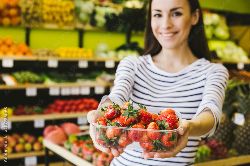 Beautiful young smiling woman holding a box with fresh strawberries and showing at the camera in a supermarket