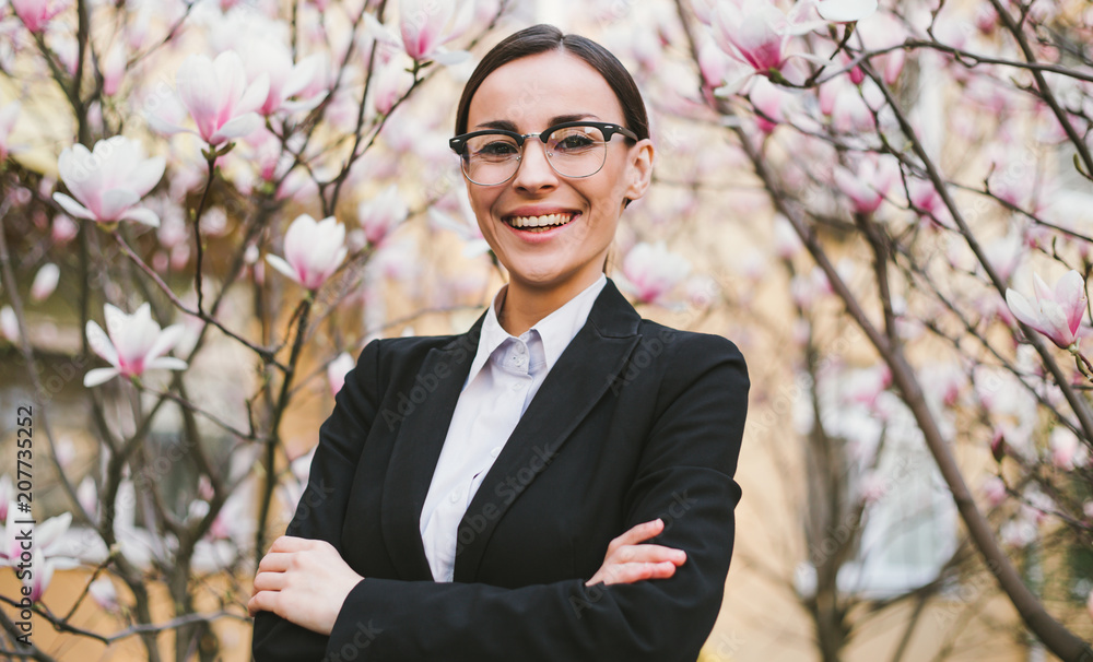 Smiling beautiful business woman in glasses and a black suit against the background of blooming magnolias. Smiling beautiful business woman in glasses and a black suit against the background of blooming magnolias.