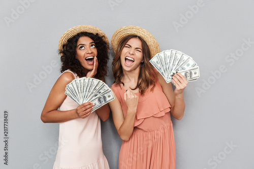 Image of two excited multiethnic women, caucasian and african american girls wearing summer clothing holding lots of cash money dollar bills, isolated over gray background