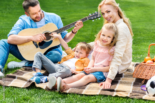 happy family playing guitar and sitting together on plaid at picnic