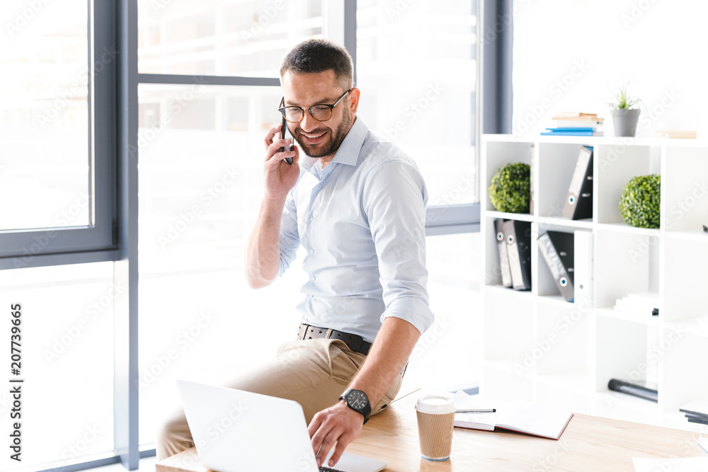Image of modern man in formal wear talking on black smartphone about business, while sitting on table in office