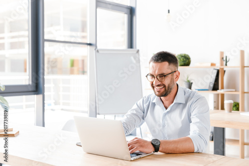 Photo of joyous office man 30s in white shirt sitting at desk and working on laptop in business centre