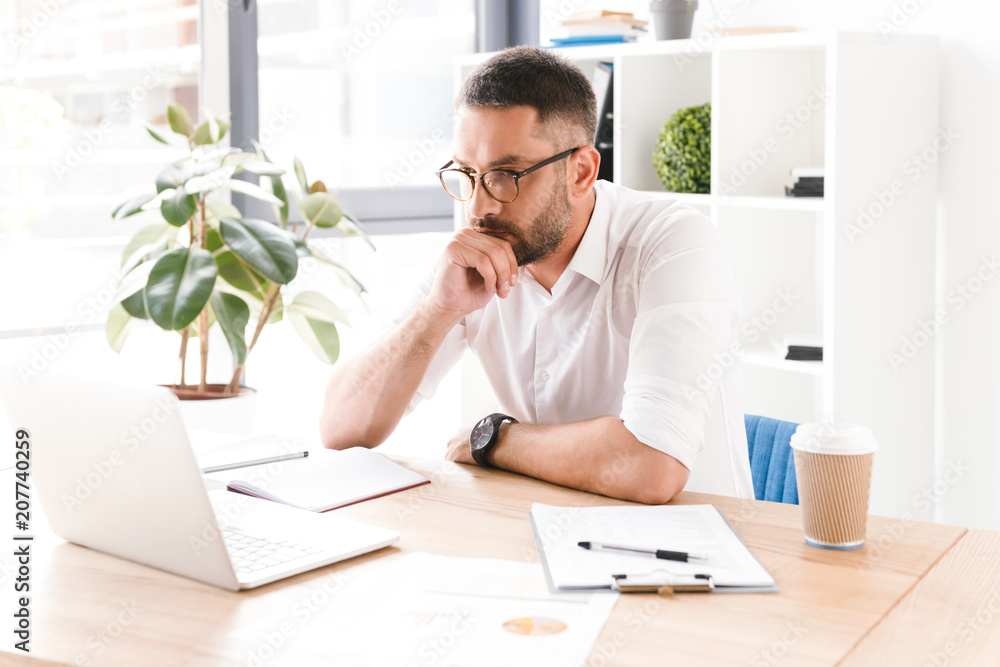 Image of concentrated confident businessman 30s wearing white shirt and glasses looking at laptop while working in office Image of concentrated confident businessman 30s wearing white shirt and glasses looking at laptop while working in office