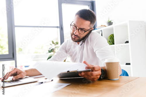 Portrait of smart businesslike man 30s in white shirt verifying information from paper documents using laptop and talking on cell phone, during work in office