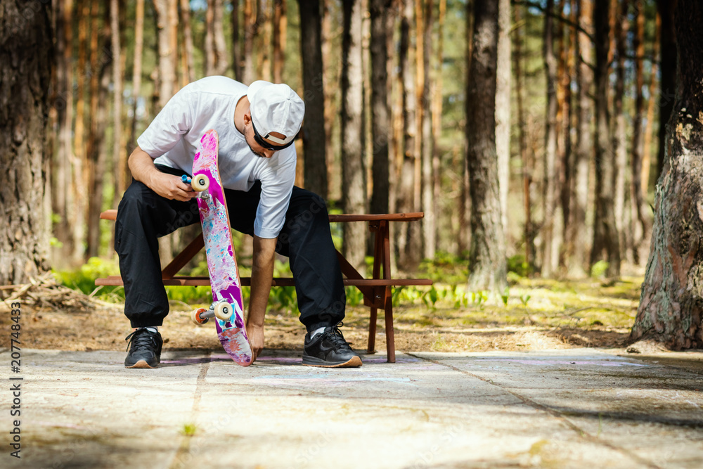 Fototapeta premium man in a white cap is sitting on a picnic table and screwing a skateboard in a summer park