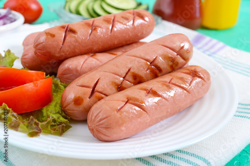 Delicious juicy grilled sausages on a plate, vegetables, ketchup and mustard on a bright background.