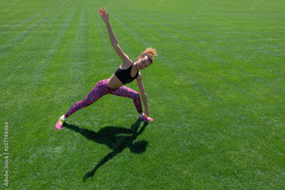 A young African-American girl in a black T-shirt, pink pants and sneakers doing sports exercises on green grass and raising her hands up. Modern style of life. Copy space. A young African-American girl in a black T-shirt, pink pants and sneakers doing sports exercises on green grass and raising her hands up. Modern style of life. Copy space.