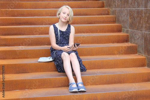 Stylish preteen girl using a phone sitting on a rusty staircase in a modern building. Child and gadget concept. 