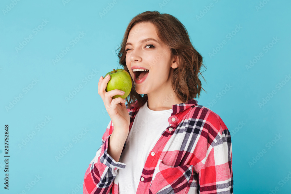 Pretty lady in plaid shirt eating green apple and happily winking looking in camera over colorful blue background Pretty lady in plaid shirt eating green apple and happily winking looking in camera over colorful blue background
