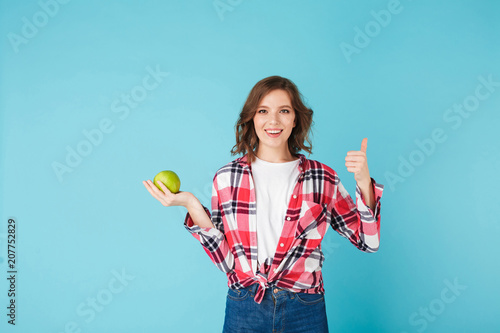 Smiling girl wearing plaid shirt and jeans and holding green apple while happily looking in camera and showing thumb up gesture over colorful blue background © Anton