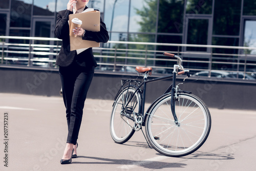 cropped shot of businesswoman with documents and coffee to go talking on smartphone while walking on street with bicycle parked behind