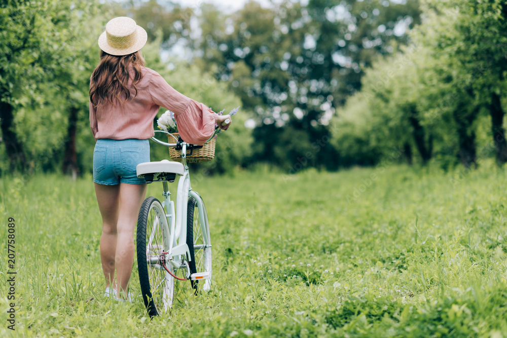 back view of young woman with retro bicycle with wicker basket full of flowers in forest back view of young woman with retro bicycle with wicker basket full of flowers in forest