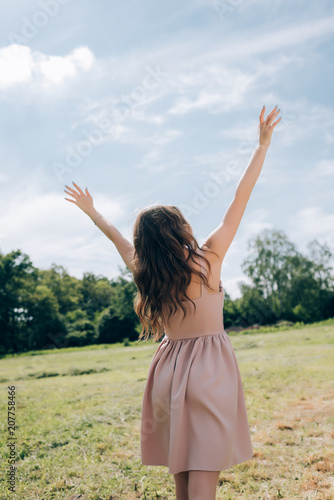 rear view of woman in stylish dress with outstretched arms standing in meadow with blue sky on background