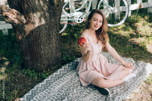 smiling attractive woman with ripe apple in hand resting on blanket under tree at countryside