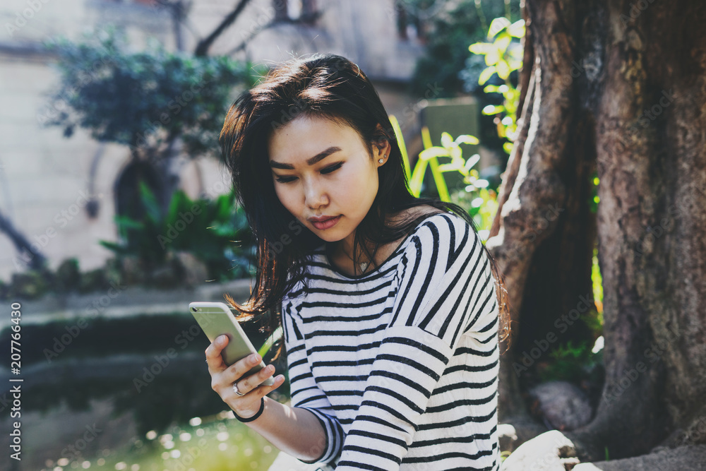 Young asian woman in casual outfit watching videos on a mobile phone while sitting in city park. Hipster girl chatting online by a mobile phone connected to free city wifi connection. Young asian woman in casual outfit watching videos on a mobile phone while sitting in city park. Hipster girl chatting online by a mobile phone connected to free city wifi connection.