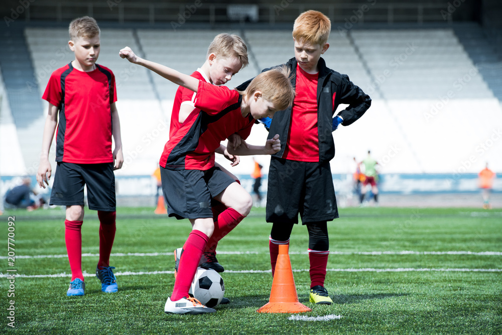 Full length portrait of junior football team practicing in stadium, focus on boy leading ball round orange cone, copy space