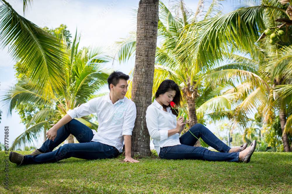 Young happy lovely couple sitting in the green park. Under the coconut tree. Man giving red roses to Young women.