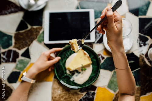 Top view of woman's hand holding the spoon with peace of pie. View from above of modern mosaic table with tablet, plate with cake, coffe.