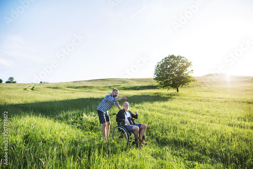 An adult hipster son with senior father in wheelchair on a walk in nature at sunset.