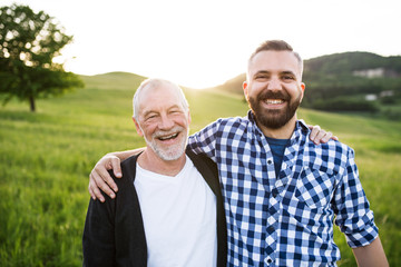 A portrait of an adult hipster son with senior father in nature at sunset, arms around each other.