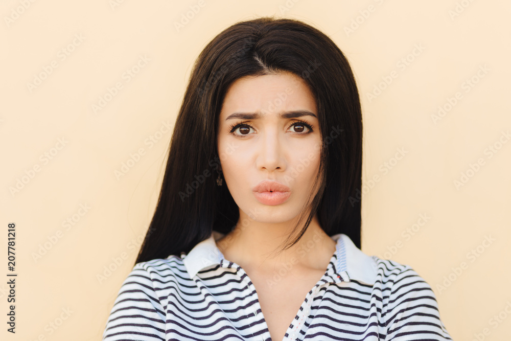 Gloomy brunette young female pouts lips in discontent, being dissatisfied with something, has healthy pure skin, dark eyes and make up, dressed in casual clothes, isolated over studio background Gloomy brunette young female pouts lips in discontent, being dissatisfied with something, has healthy pure skin, dark eyes and make up, dressed in casual clothes, isolated over studio background