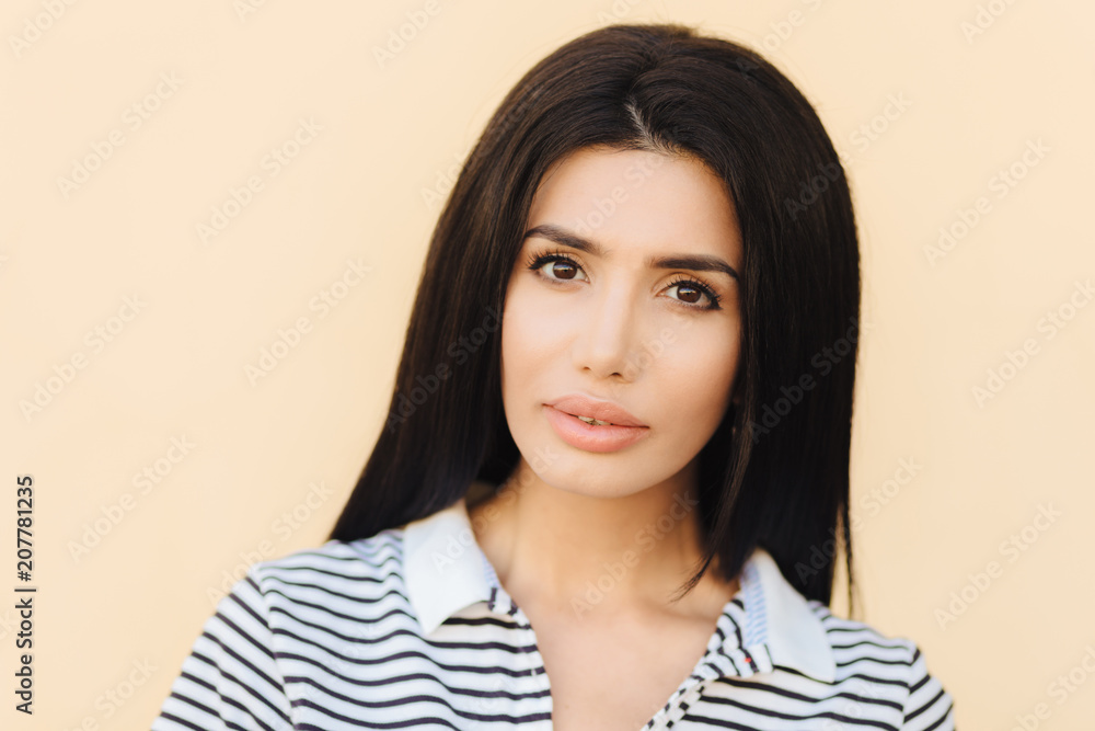Serious beautiful brunette female looks confidently into camera, has long straight hair, listens attentively interlocutor, wears casual striped t shirt, isolated on light background. People and beauty Serious beautiful brunette female looks confidently into camera, has long straight hair, listens attentively interlocutor, wears casual striped t shirt, isolated on light background. People and beauty