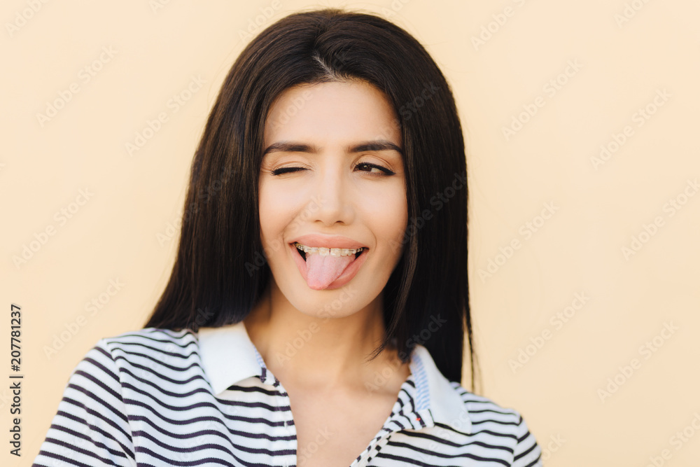 Funny joyful brunette female shows tongue, blinks with eye, makes grimace, wears braces, has long straight dark luxurious hair, dressed casually, isolated over light background. People and happiness Funny joyful brunette female shows tongue, blinks with eye, makes grimace, wears braces, has long straight dark luxurious hair, dressed casually, isolated over light background. People and happiness