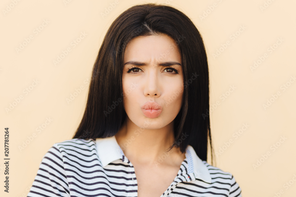 People, beauty and facial expressions concept. Discontent brunette cute woman pouts lips and frowns face, has sullen look, dressed in casual striped t shirt, has gloomy expression after quarrel People, beauty and facial expressions concept. Discontent brunette cute woman pouts lips and frowns face, has sullen look, dressed in casual striped t shirt, has gloomy expression after quarrel