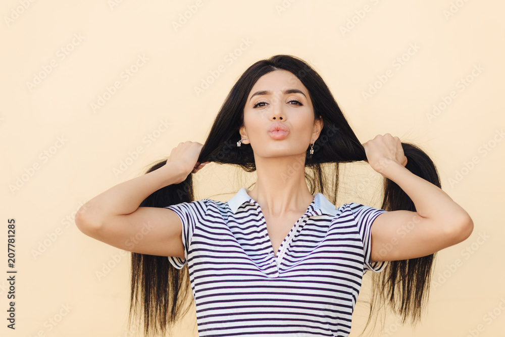 Lovely young Caucasian female shows her long straight dark hair, pouts lips, demonstrates natural beauty, poses against light background, dressed in striped t shirt. Hair care and beauty concept Lovely young Caucasian female shows her long straight dark hair, pouts lips, demonstrates natural beauty, poses against light background, dressed in striped t shirt. Hair care and beauty concept