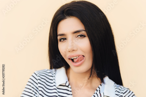 Portrait of attractive female model licks mouth with tongue, has curious look, healthy skin and dark hair, cunning expression, isolated over studio wall. Facial expressions and beauty concept © VK Studio