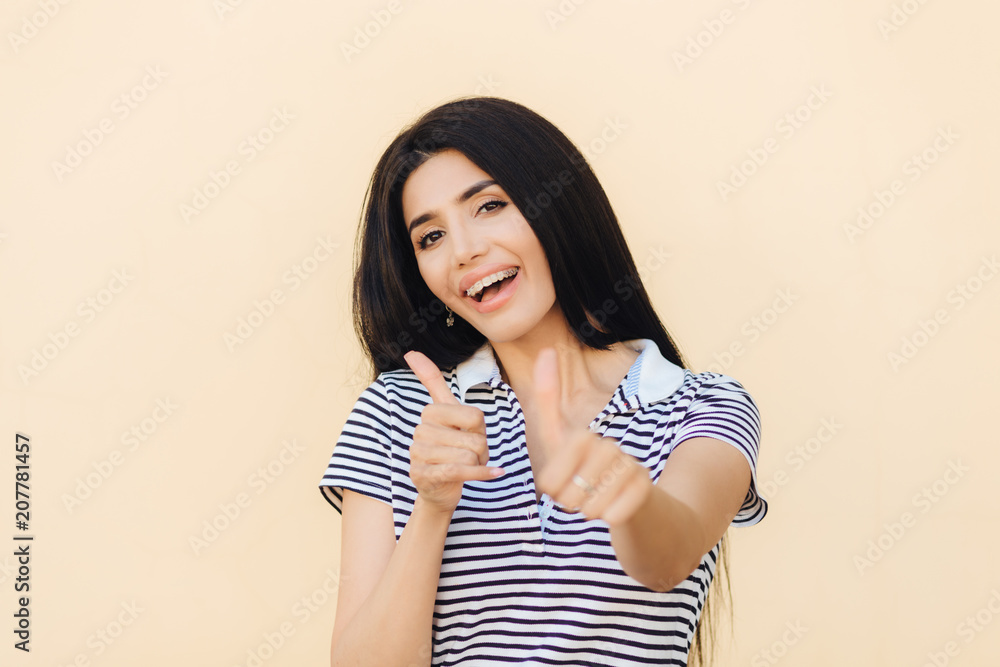 Shot of attractive European female gives double thumb up, has positive expression, has white teeth with brackets, dressed casually, makes okay and approval gesture. People and body language. Shot of attractive European female gives double thumb up, has positive expression, has white teeth with brackets, dressed casually, makes okay and approval gesture. People and body language.