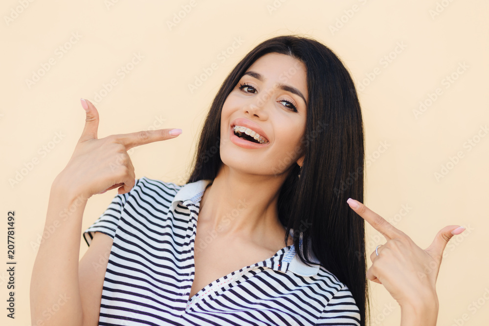 Portrait of happy gorgeous female model indicates with fore fingers at her face, advertises braces for even teeth, has dark long straight hair, shining eyes and perfect healthy skin, models indoor Portrait of happy gorgeous female model indicates with fore fingers at her face, advertises braces for even teeth, has dark long straight hair, shining eyes and perfect healthy skin, models indoor