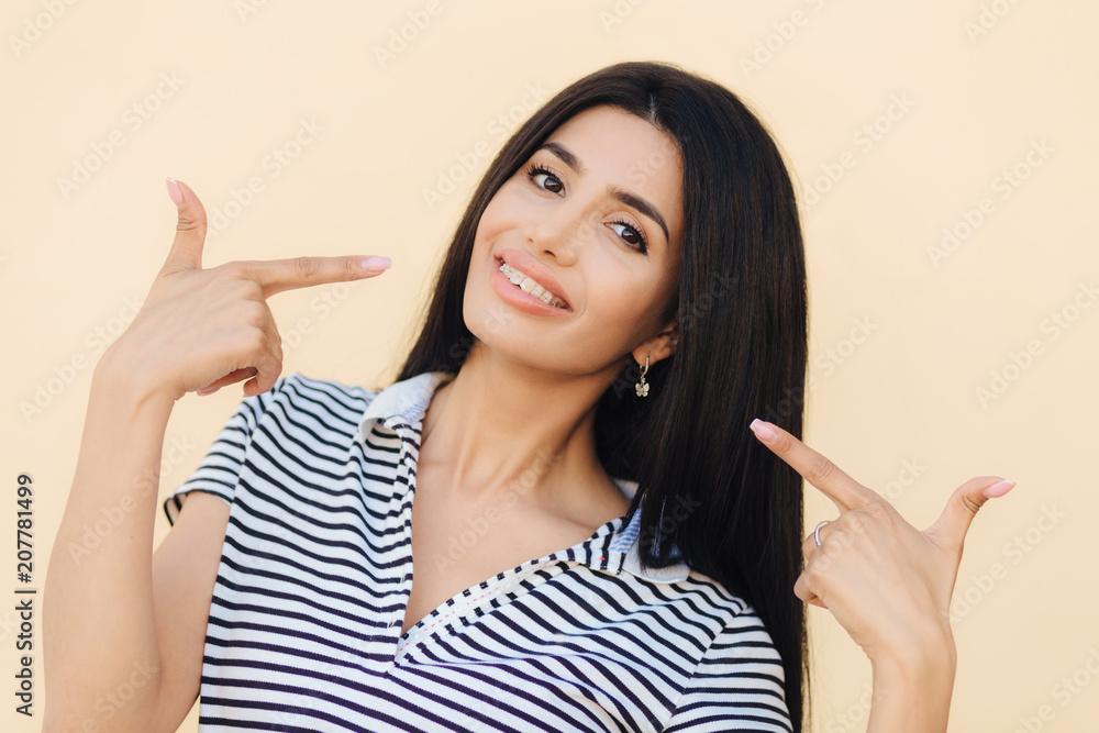 Horizontal portrait of happy brunette female model has broad smile, white teeth, indicates with both index fingers at mouth, cares about her beauty, isolated over light pink studio background