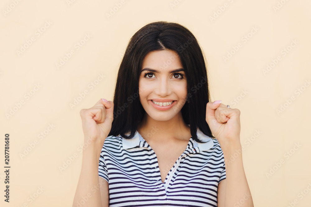 Headshot of happy brunette female model clenches fists, hopes for something good, has broad smile with white perfect teeth, celebrates success, isolated over beige studio wall. I will do it! Headshot of happy brunette female model clenches fists, hopes for something good, has broad smile with white perfect teeth, celebrates success, isolated over beige studio wall. I will do it!