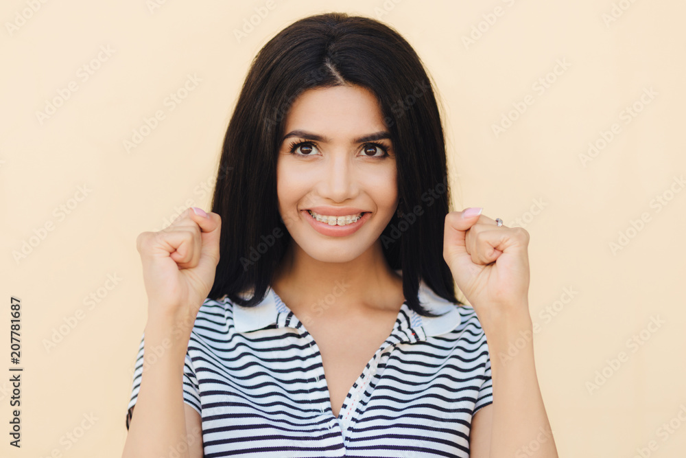 Candid shot of cheerful brunette young female has big hope, keeps hands in fists, believes in better, stands against beige background, has pleasant smile on face. People and happiness concept Candid shot of cheerful brunette young female has big hope, keeps hands in fists, believes in better, stands against beige background, has pleasant smile on face. People and happiness concept