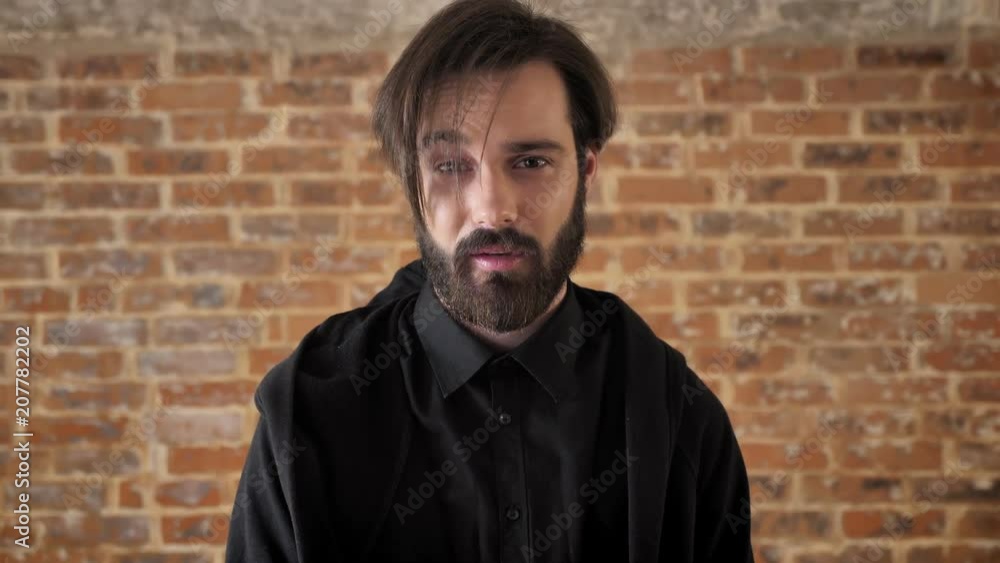 Young indifferent guy raises eyebrow, watching at camera, brick background Young indifferent guy raises eyebrow, watching at camera, brick background