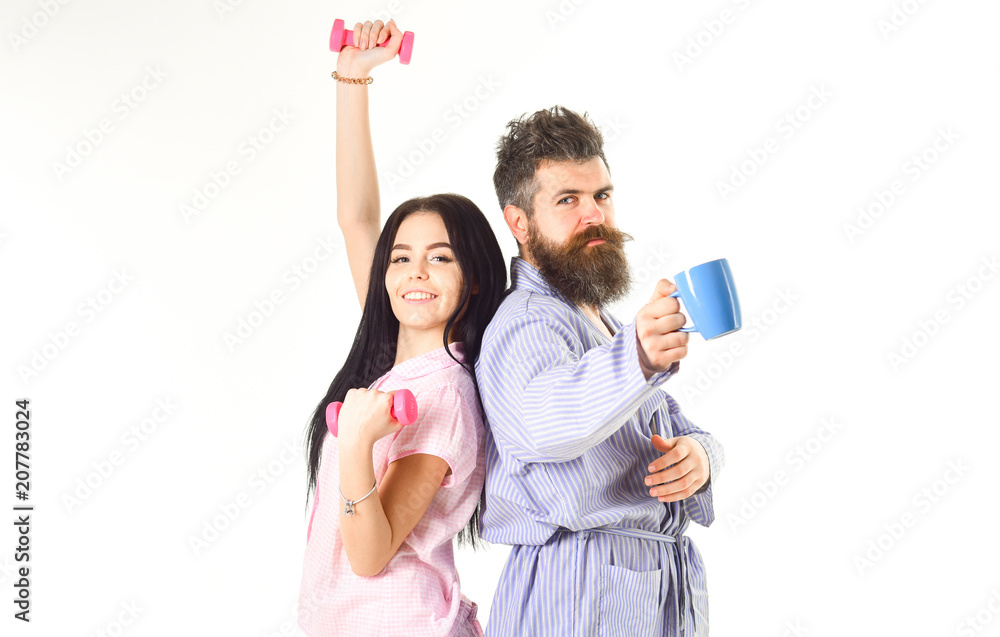 Couple in love in pajama, bathrobe stand isolated on white background. Morning alternative concept. Girl with dumbbell, man with coffee cup. Couple, family on sleepy faces, full of energy.