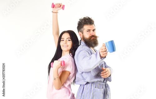 Couple in love in pajama  bathrobe stand isolated on white background. Morning alternative concept. Girl with dumbbell  man with coffee cup. Couple  family on sleepy faces  full of energy.