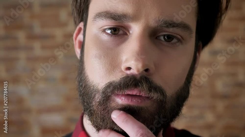 Young attractive serious man is touching his beard, watching at camera, brick background photo