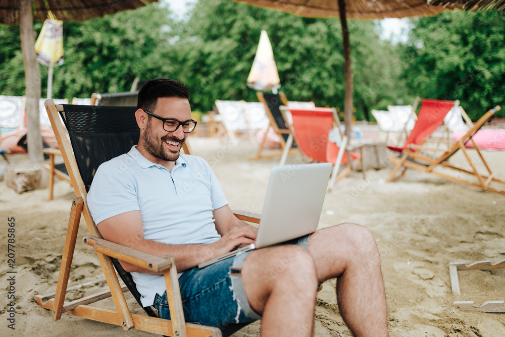 Young smiling man using laptop at the beach.