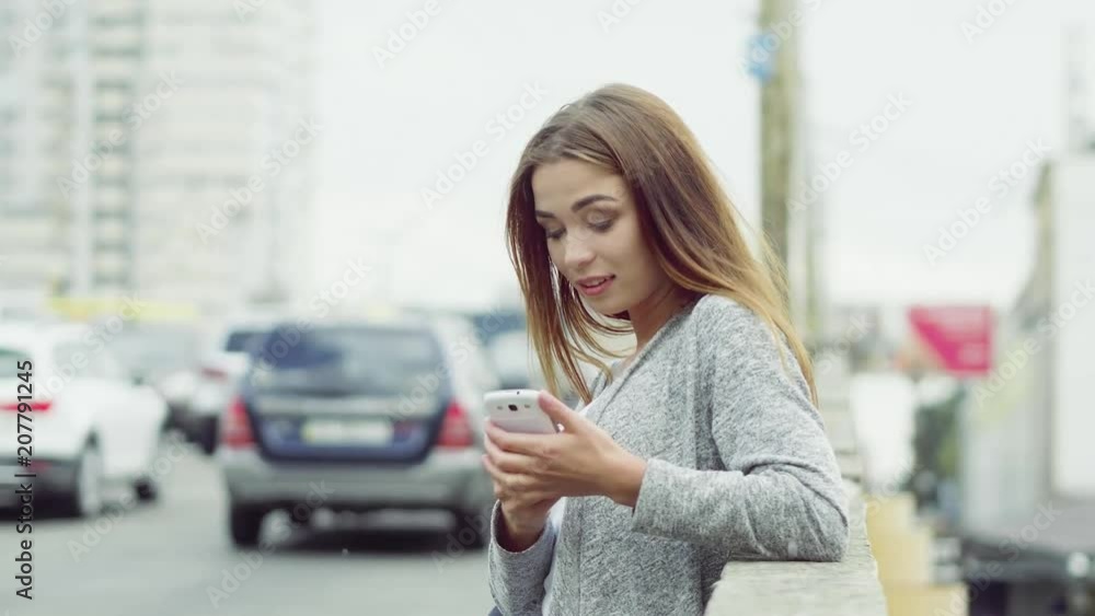 Smiling charming woman reading sms from friend while standing near traffic way. Big city concept. Smiling charming woman reading sms from friend while standing near traffic way. Big city concept.