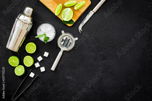 Make mojito cocktail with lime and mint. Shaker, strainer, glass near slices of lime on cutting board on black background top view copy space