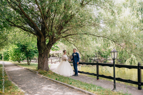 The husband and wife in wedding clothes are posing under an old broad tree with heavy branches. The newlyweds are just getting married walking in the park and holding hands.