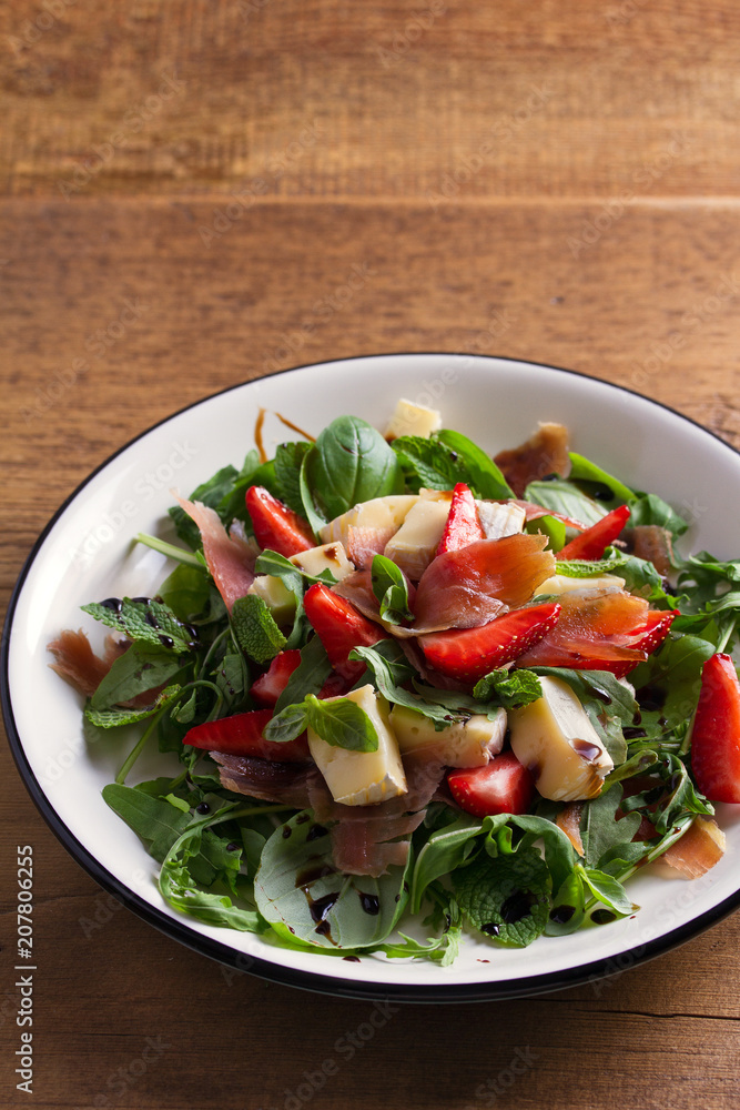 Strawberry, basil, mint, arugula salad with brie cheese and jamon on white plate on wooden table. vertical