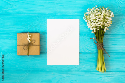 Mockup with bouquet of flowers lily of the valley and empty paper sheet on blue wooden table, gift box top view,