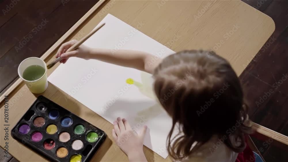 Top view of a little girl with long fair hair sitting at a wooden table for kids and drawing. Concept of creativity. Right to left pan slow motion medium shot Top view of a little girl with long fair hair sitting at a wooden table for kids and drawing. Concept of creativity. Right to left pan slow motion medium shot