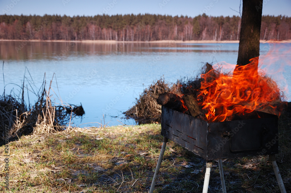 The fire burns in the grill against the backdrop of a beautiful lake.
