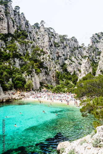 View of the calanque of En-Vau, a natural creek with crystal clear water and white sandy beach between Marseille and Cassis, part of the Calanques National Park, with people sunbathing and swimming.