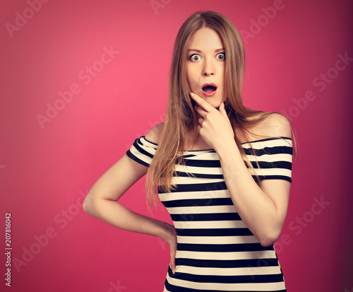 Fun strict surprising woman in striped dress holding hand near the mouth with big opened eyes on pink background and looking serious. Toned closeup portrait